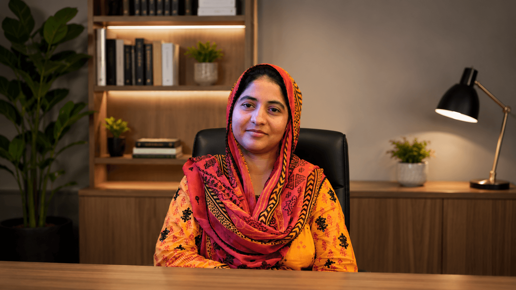 Smiling woman in a vibrant orange and red headscarf sitting at a wooden desk.